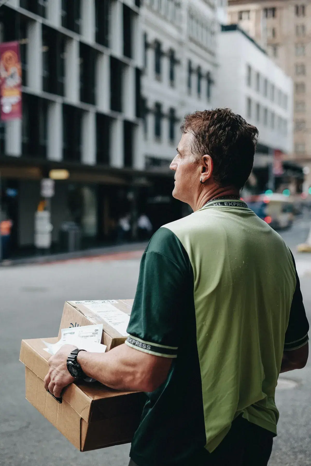 A delivery courier in a green shirt carrying a cardboard box down a city street