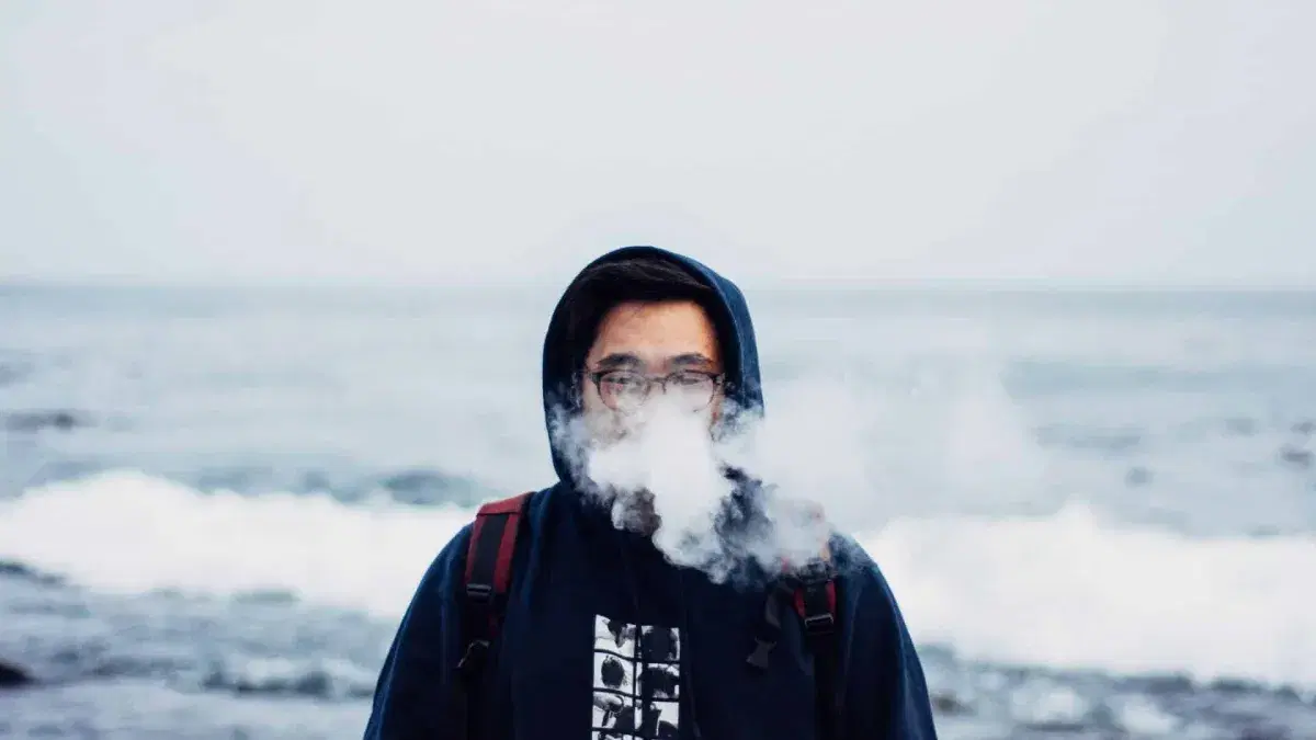 a person in a hoodie exhaling vapour by the sea